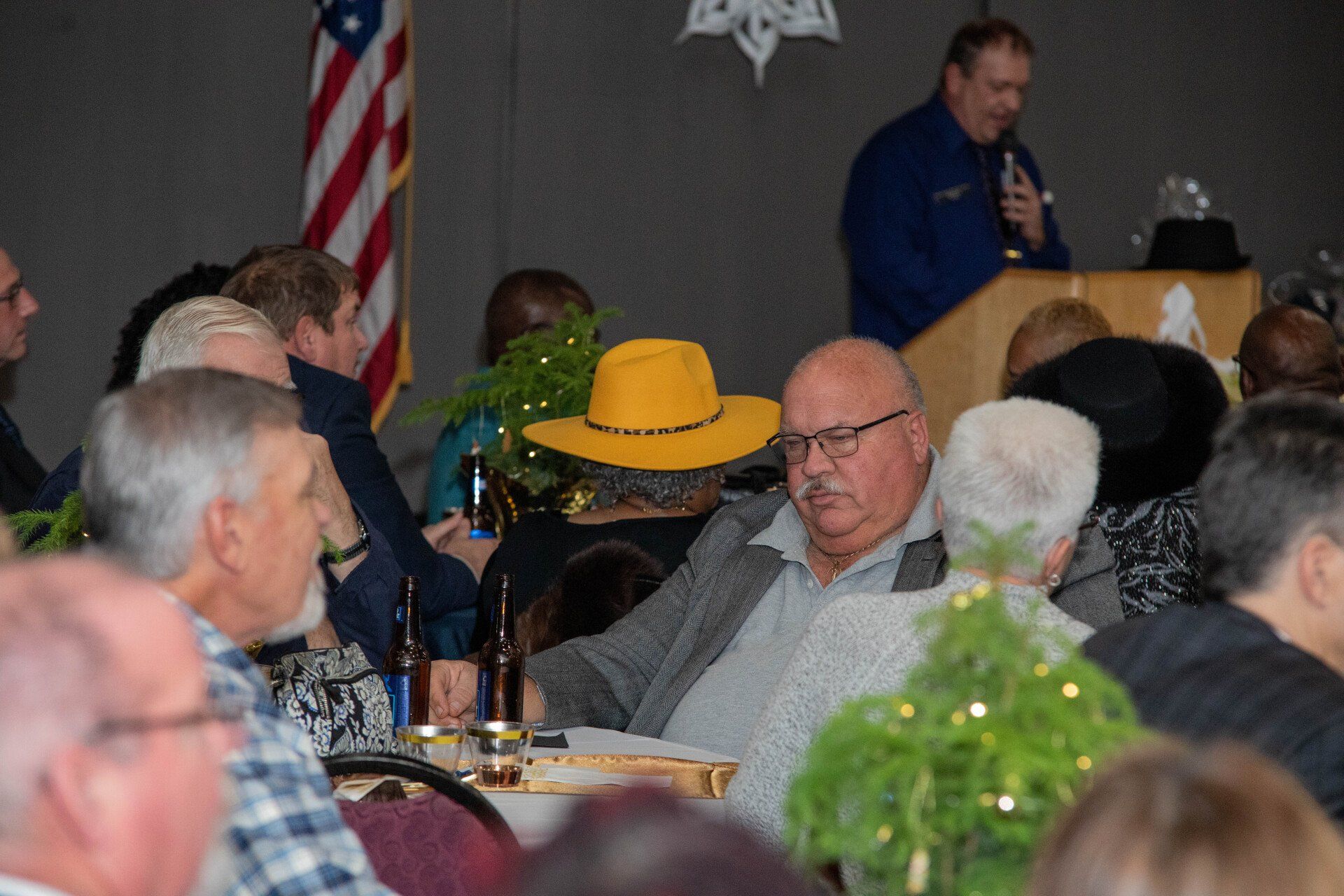 A man speaks at a podium in a crowded room. Guests sit at tables decorated with greenery. An American flag hangs in the background.