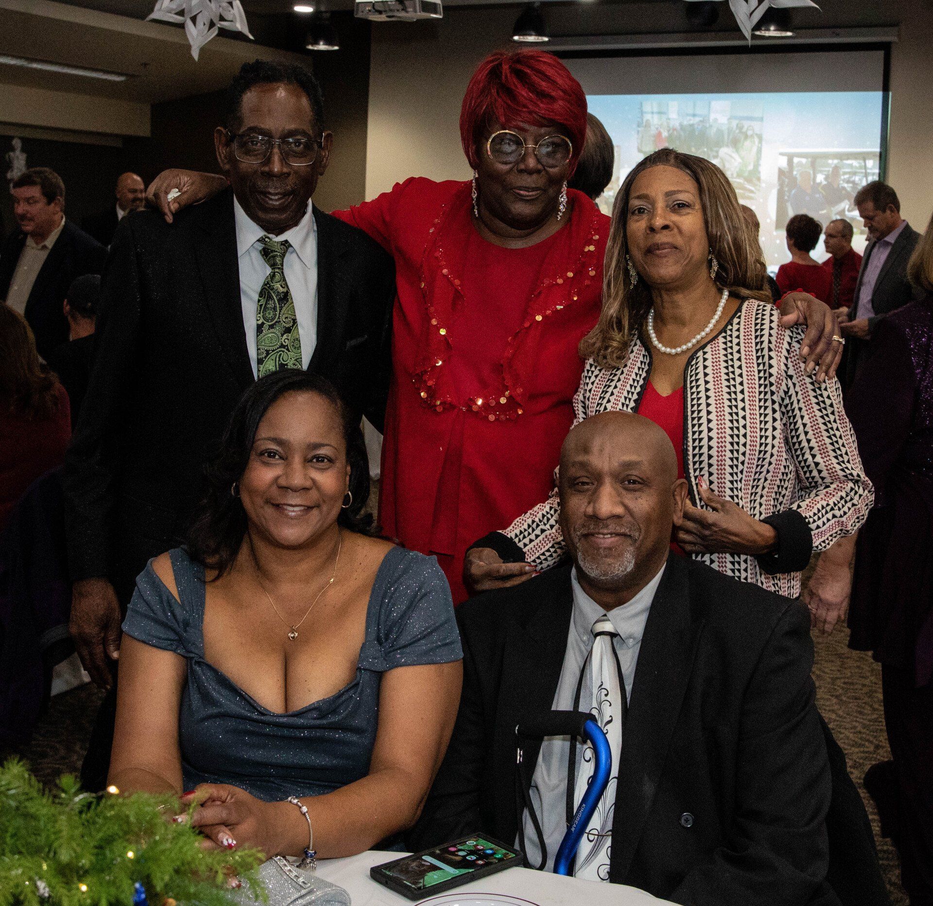 Group photo of six Black people at an event. People smile, some dressed in formal attire.  A man uses a cane.  Background includes people and a screen.