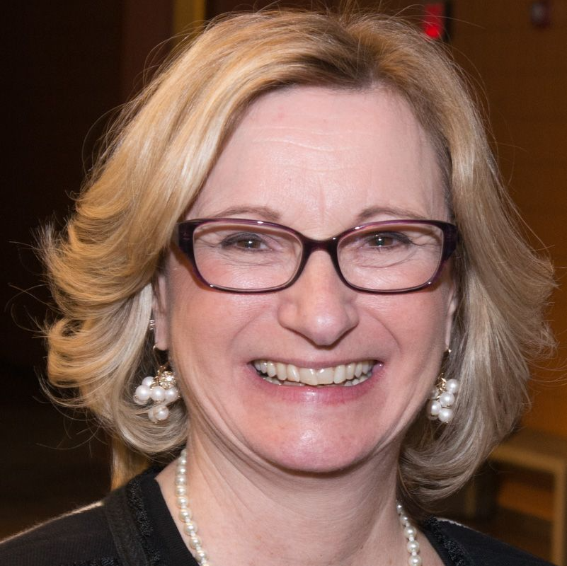 Woman with blonde hair, glasses, and pearl earrings smiles. She wears a pearl necklace and a black top.