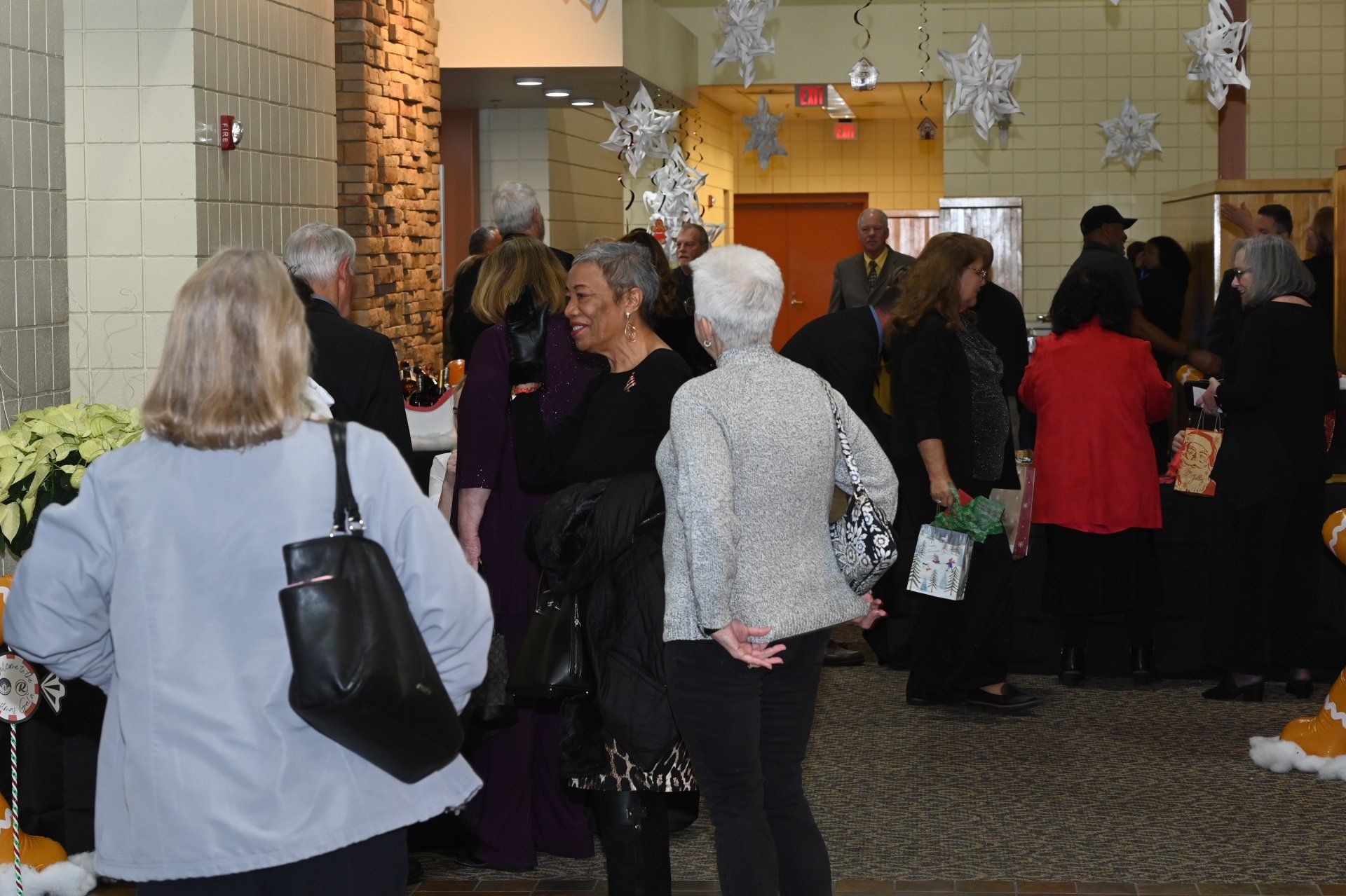 People gather in a decorated indoor space; some are talking, others browsing. Decorations include hanging snowflakes and poinsettias.