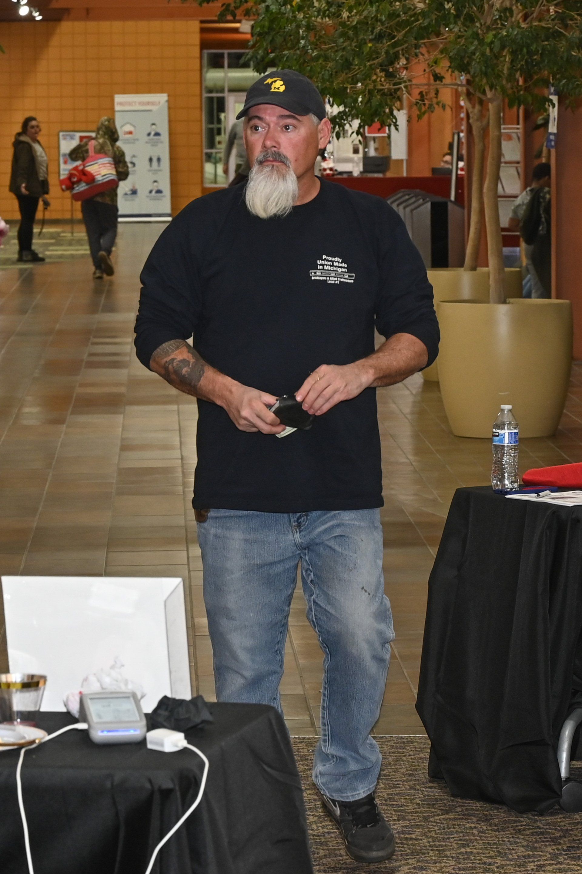 Man with a white beard stands by a table in a building. He wears a navy shirt, jeans, and a cap.