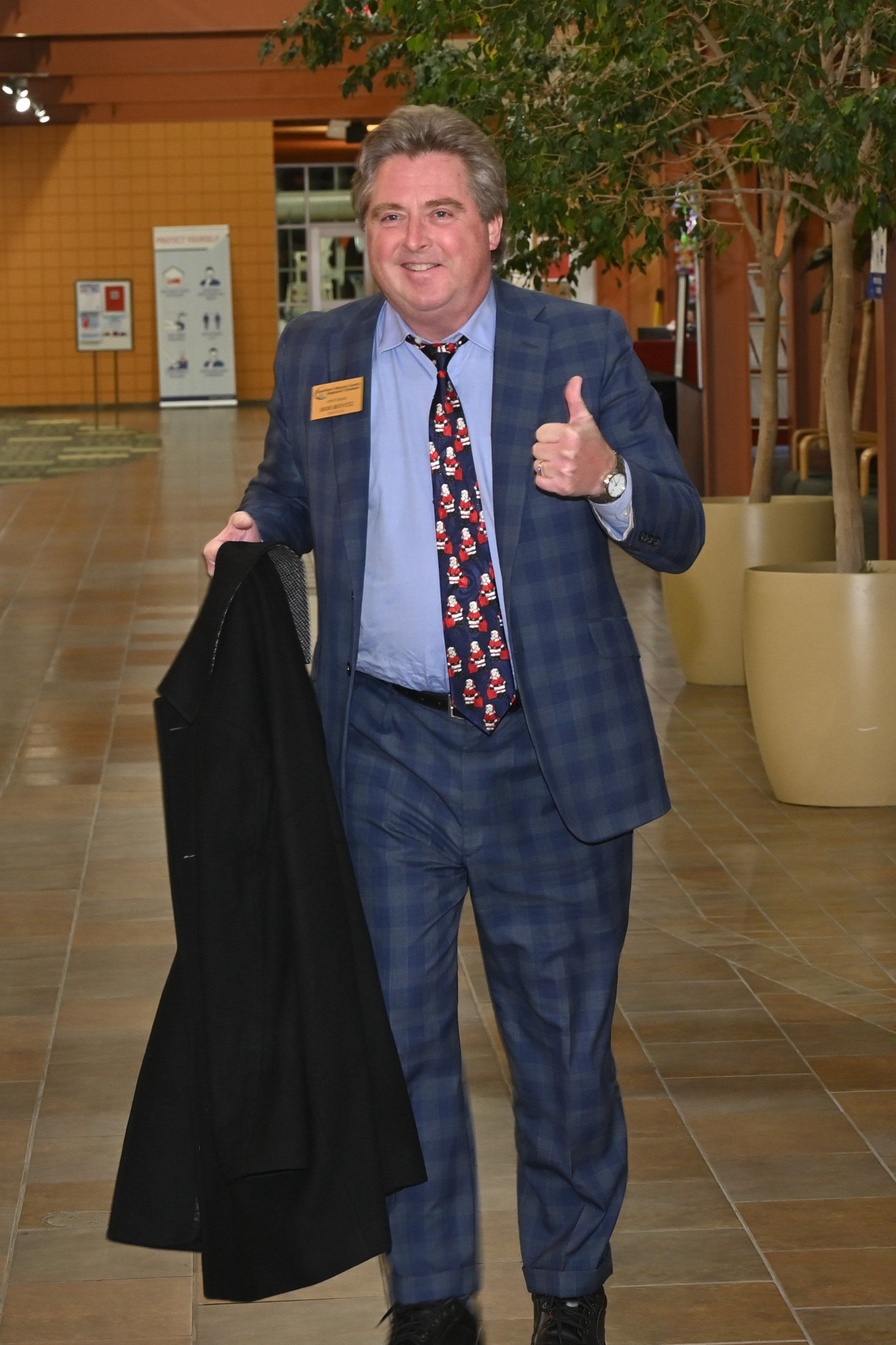 A man in a blue plaid suit gives a thumbs-up. He is holding a black coat in a hallway.