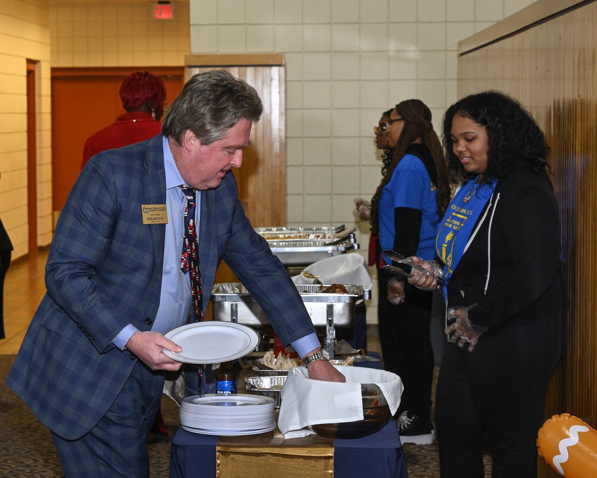 Man in suit serves food from a buffet to a woman. Several others wait in line.