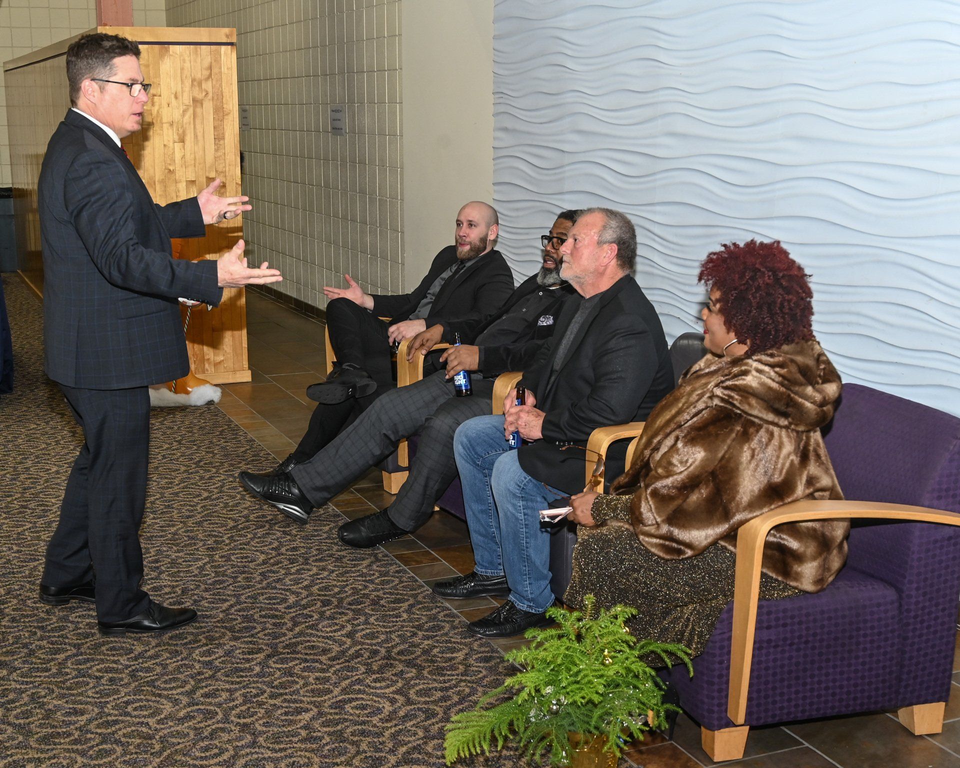 A man in a suit gestures to a group seated on a purple couch. The group includes three men and one woman, indoors.