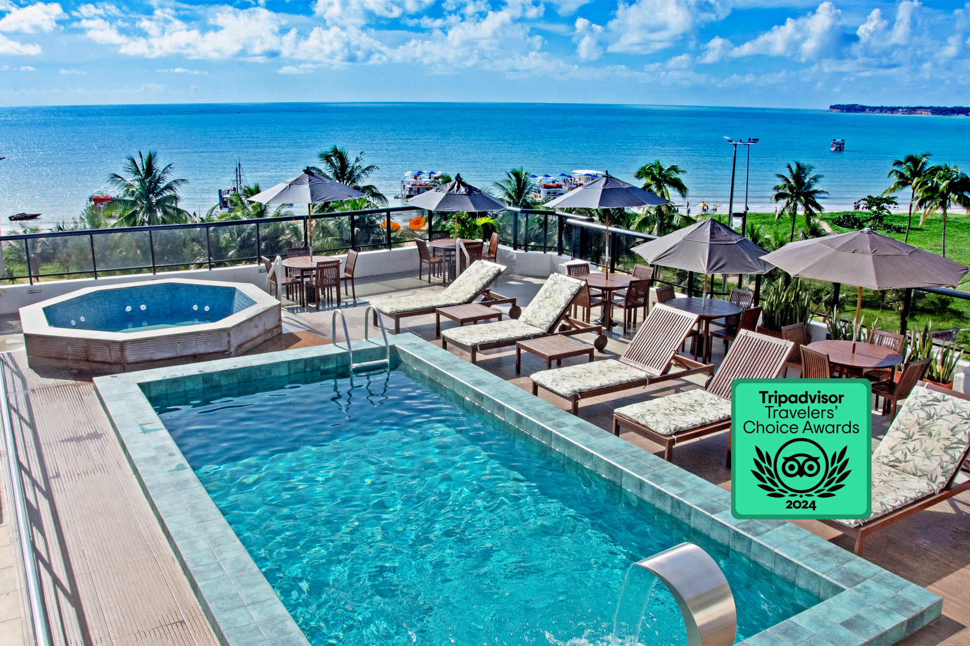 Uma piscina e jacuzzi na cobertura com vista para um oceano azul brilhante, com espreguiçadeiras e mesas sob guarda-sóis.