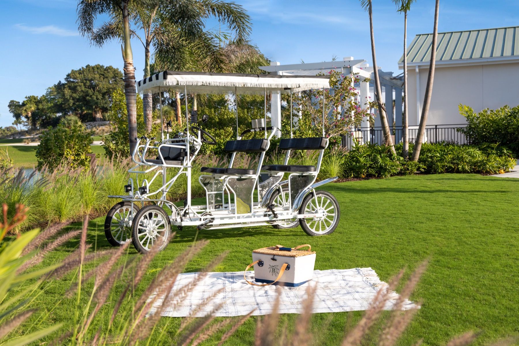 A bicycle with four seats is parked in the grass next to a picnic blanket.