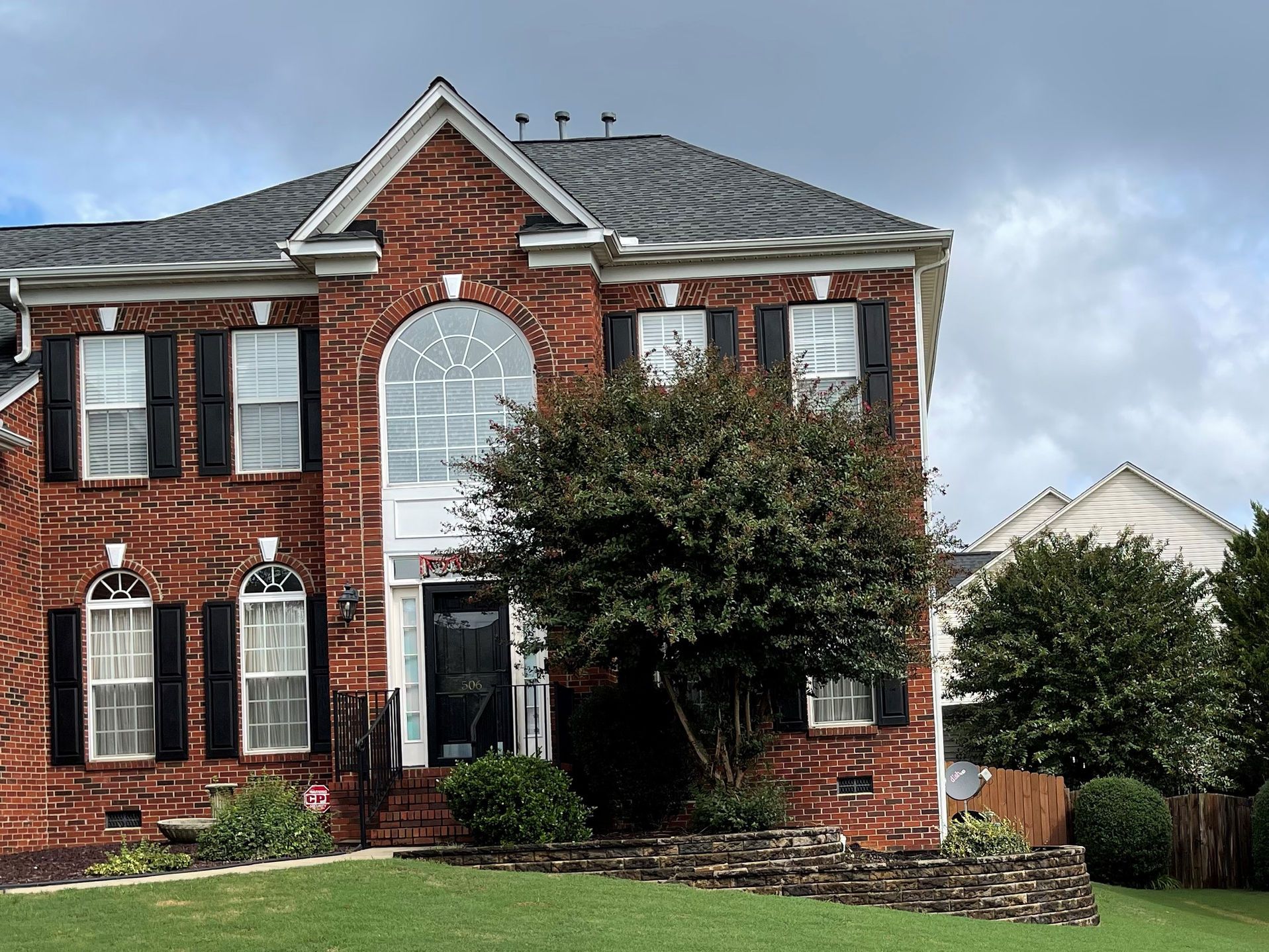 Two-story brick house with black shutters, large arched window above the front door, and a cloudy sky.