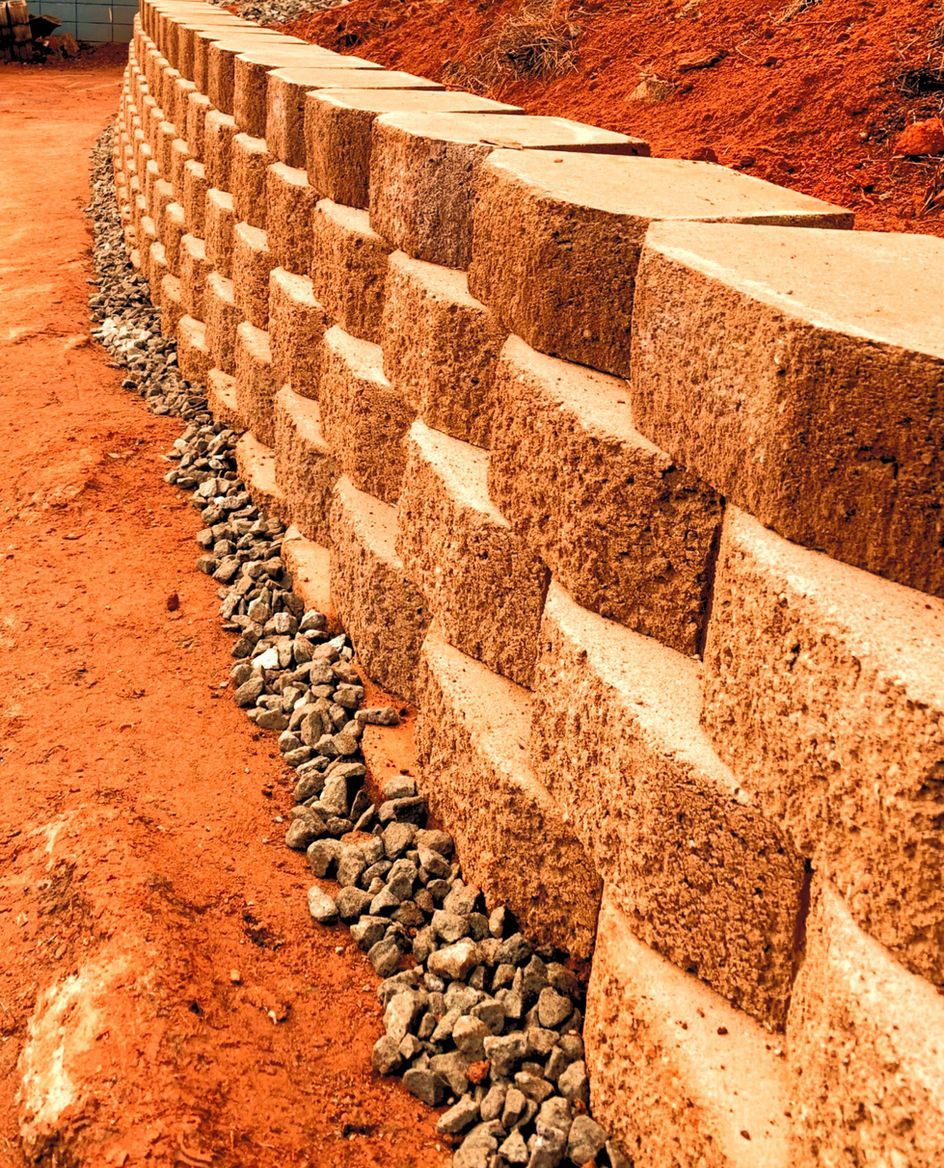 Retaining wall made of tan-colored, textured blocks on a red-brown earth slope with gravel at the base.