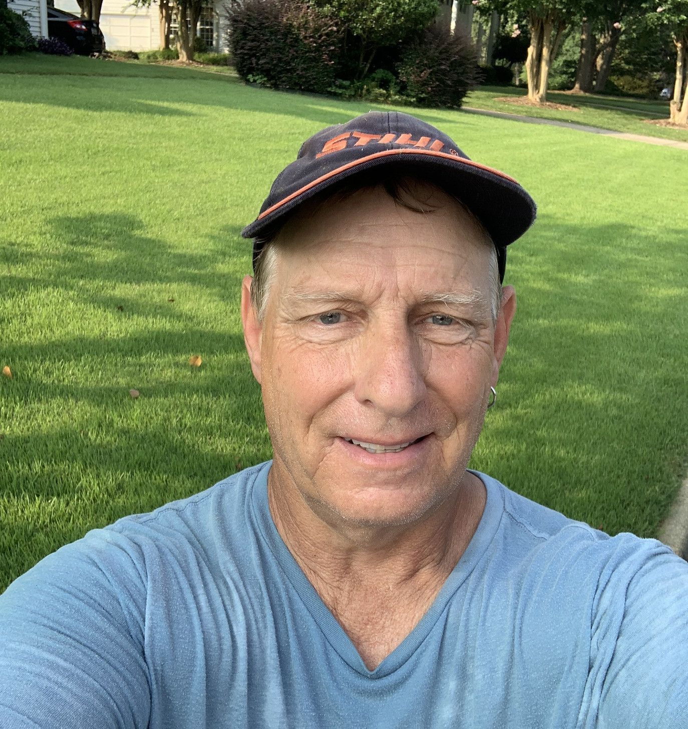 Man in a blue shirt and hat smiles outdoors on a sunny day, with green grass and trees.