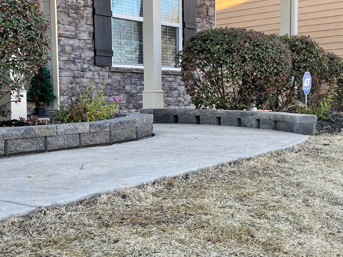 Curved concrete walkway with a low brick retaining wall, leading to a home's entrance. Dry grass and shrubs surround.