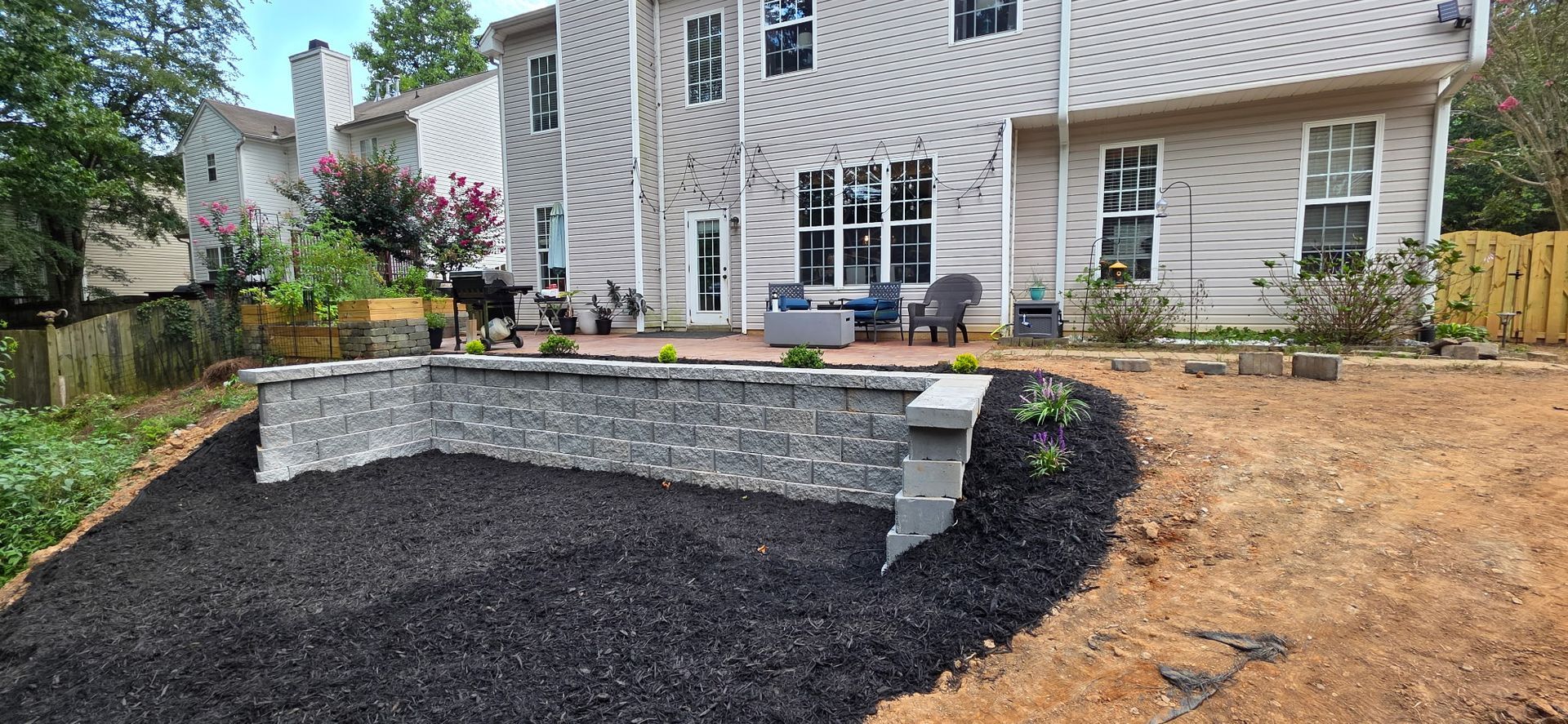 Backyard with a retaining wall, patio, and house. Black mulch in the foreground, with trees and a fence in the background.