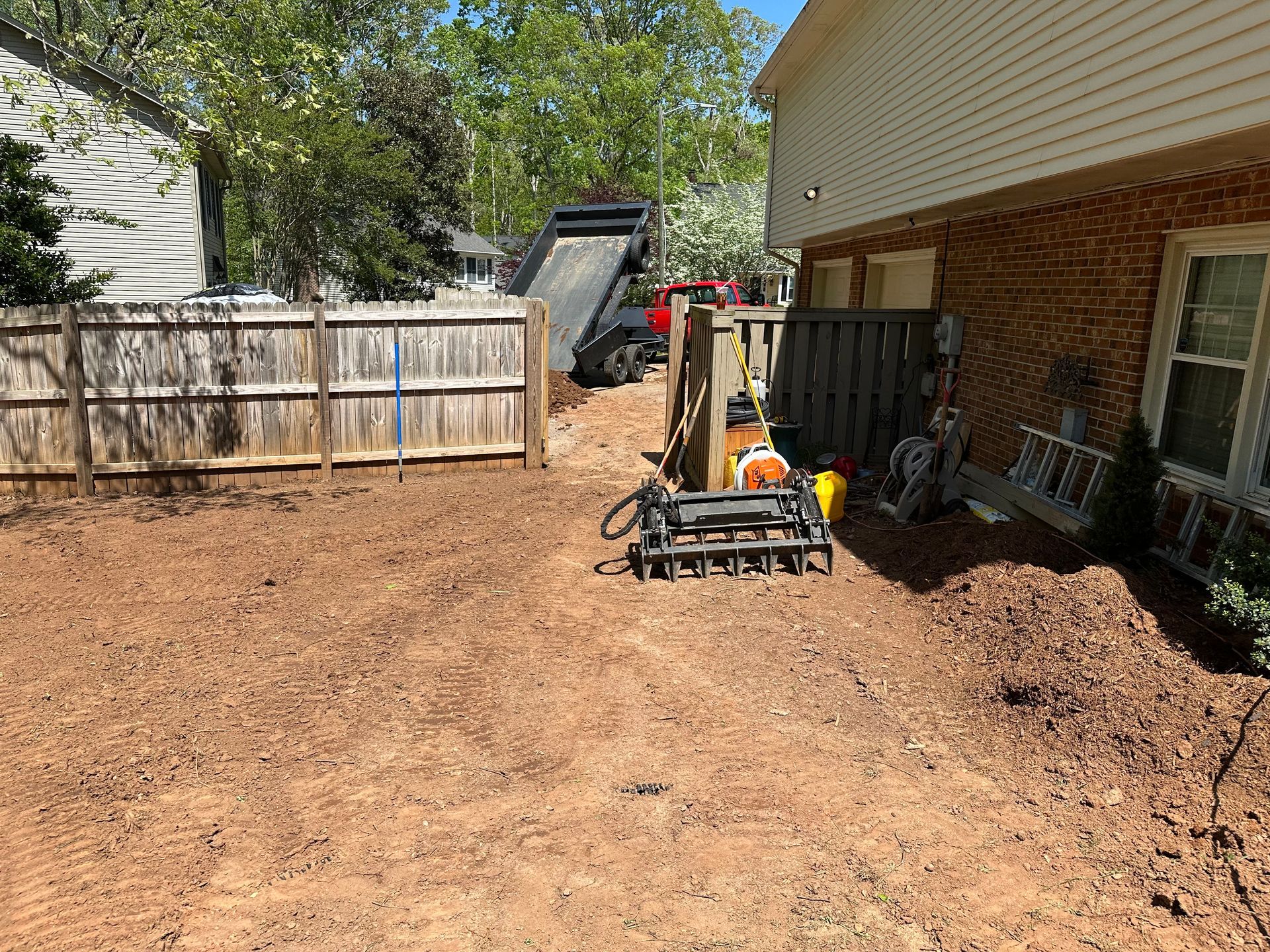 Dirt backyard with fence, house, and landscaping equipment. Dump truck in the distance.