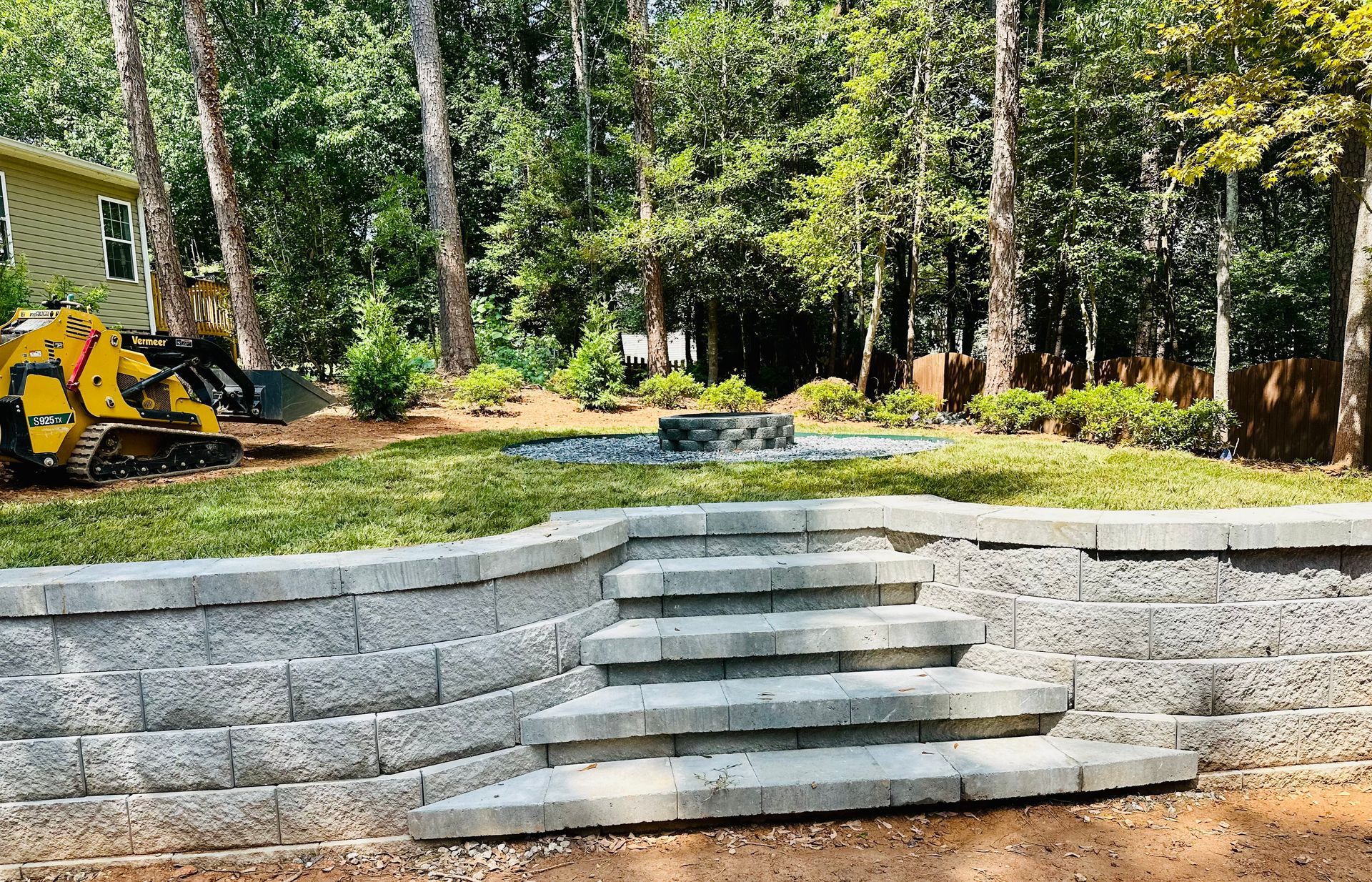 Stone steps and retaining wall leading up to a grassy backyard with a fire pit, trees, and construction equipment.