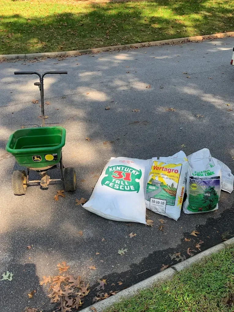 Lawn spreader next to bags of grass seed on asphalt. One bag is marked 