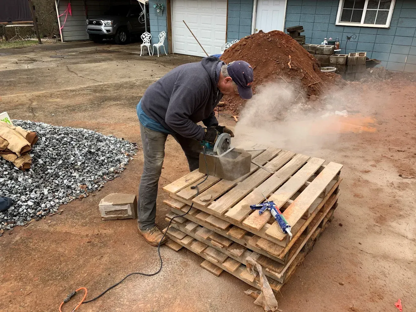 Man cuts a block on wooden pallets, creating a cloud of dust outdoors near a dirt pile and blue building.