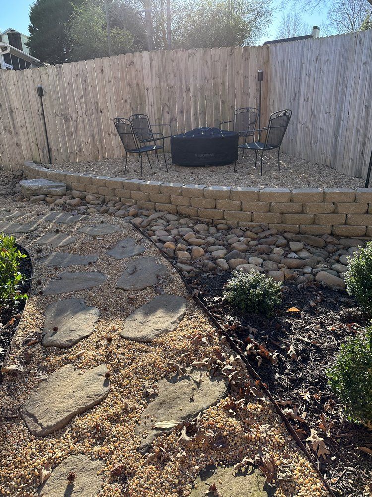 Backyard with a stone pathway, retaining wall, gravel, fire pit, and metal chairs near a wooden fence.