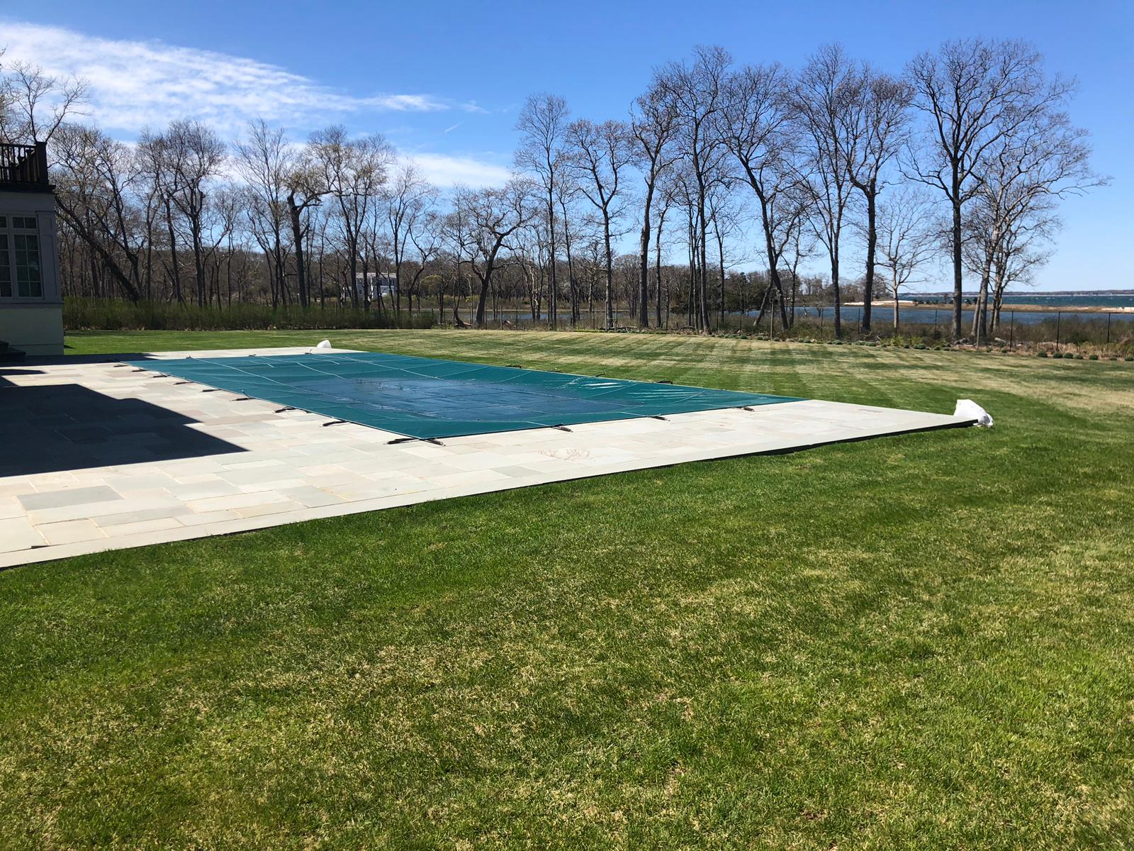 Pool covered with blue tarp, bordered by stone patio and green lawn; trees and water in the background.