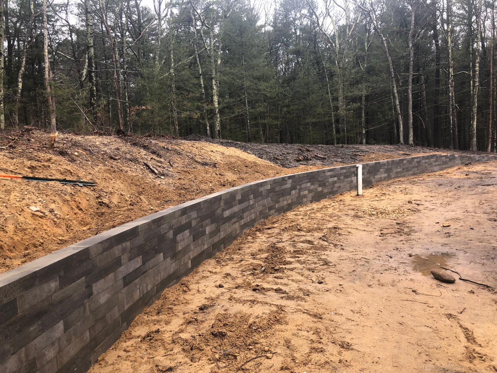 A retaining wall made of dark bricks curves along a dirt path, with trees in the background.