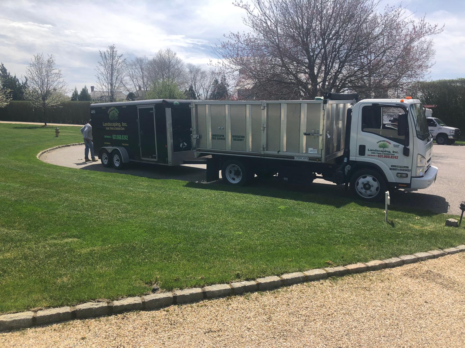 A white landscaping truck and trailer on green grass, with a person standing nearby.