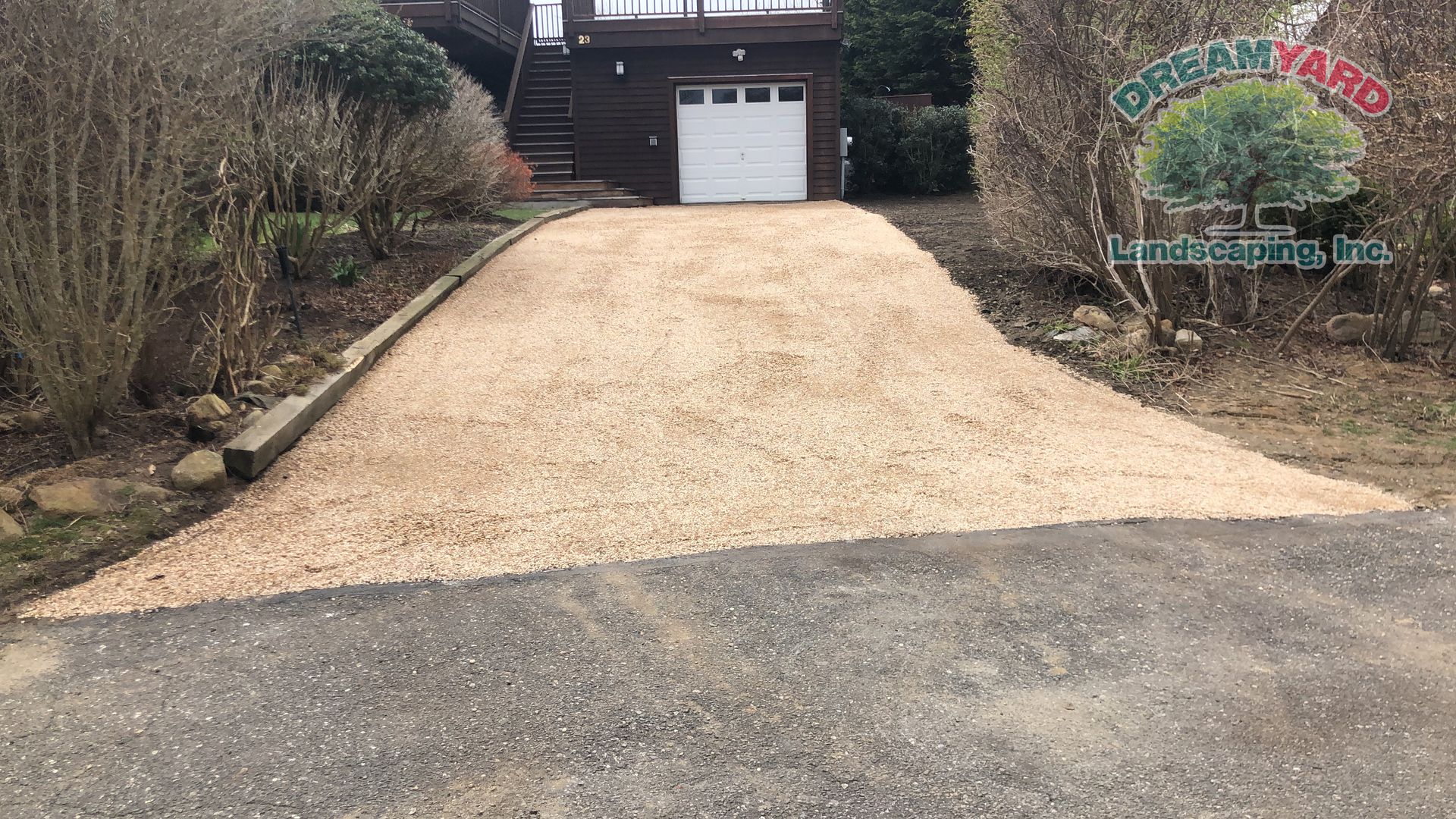 Gravel driveway leading uphill to a garage. Beige gravel, dark asphalt, and brown bushes.