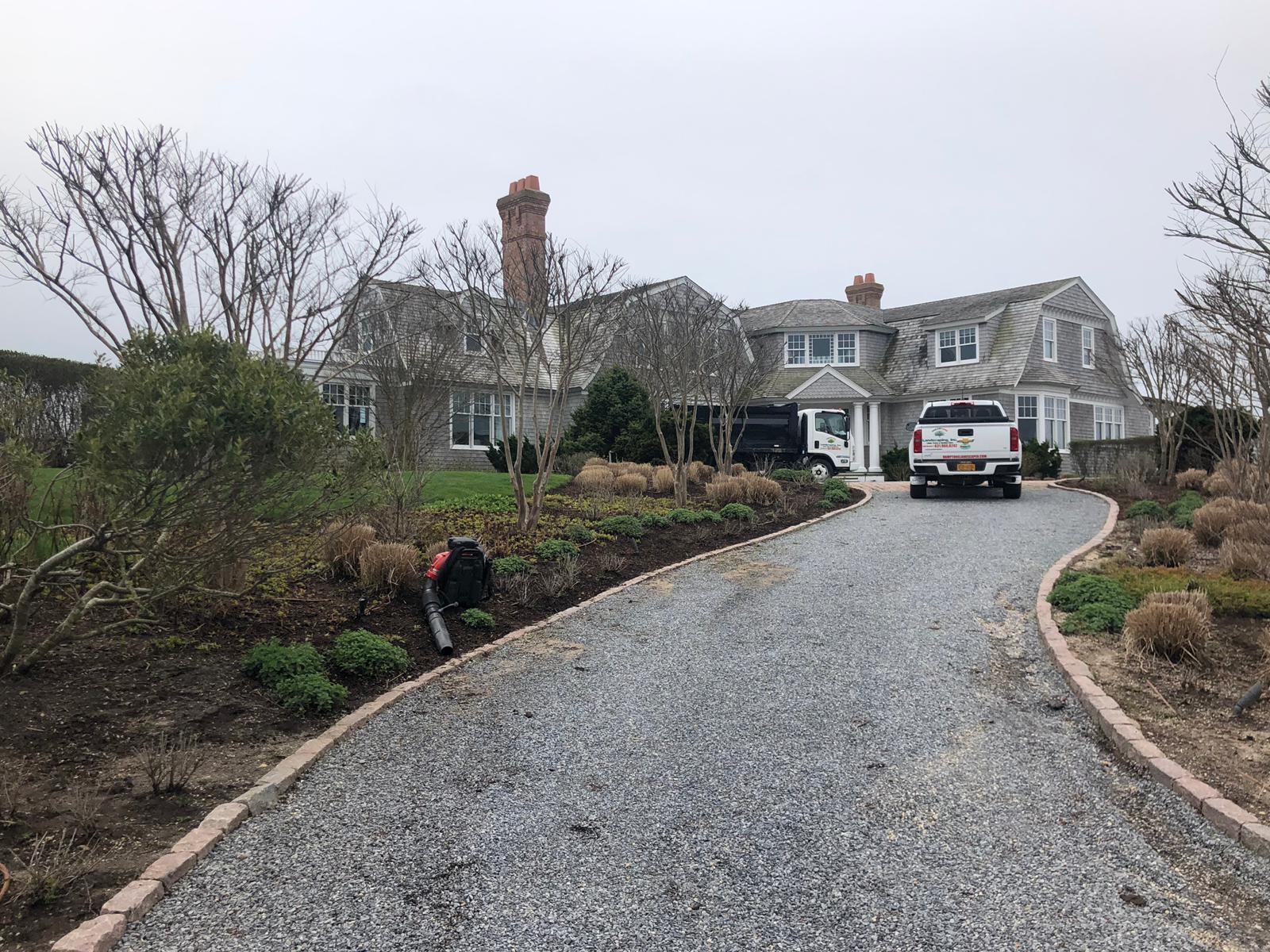 Large gray house with a gravel driveway. Landscaping and two work trucks are visible. Overcast sky.