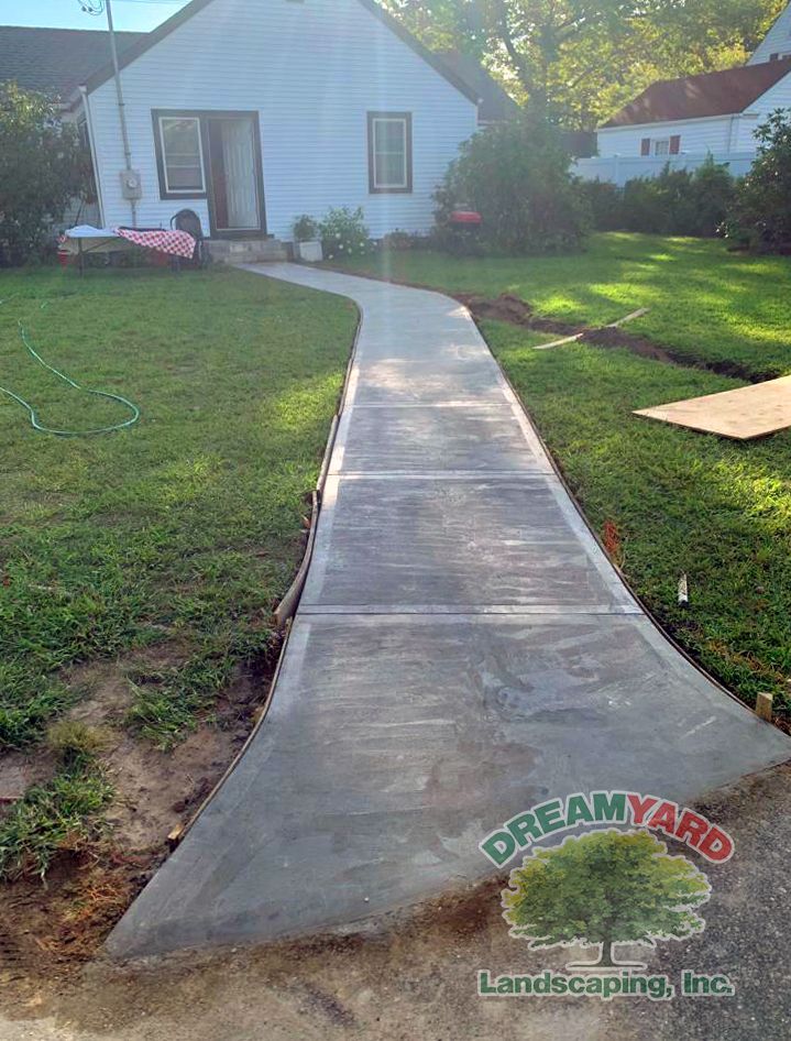 A new concrete sidewalk curves towards a white house with green grass.
