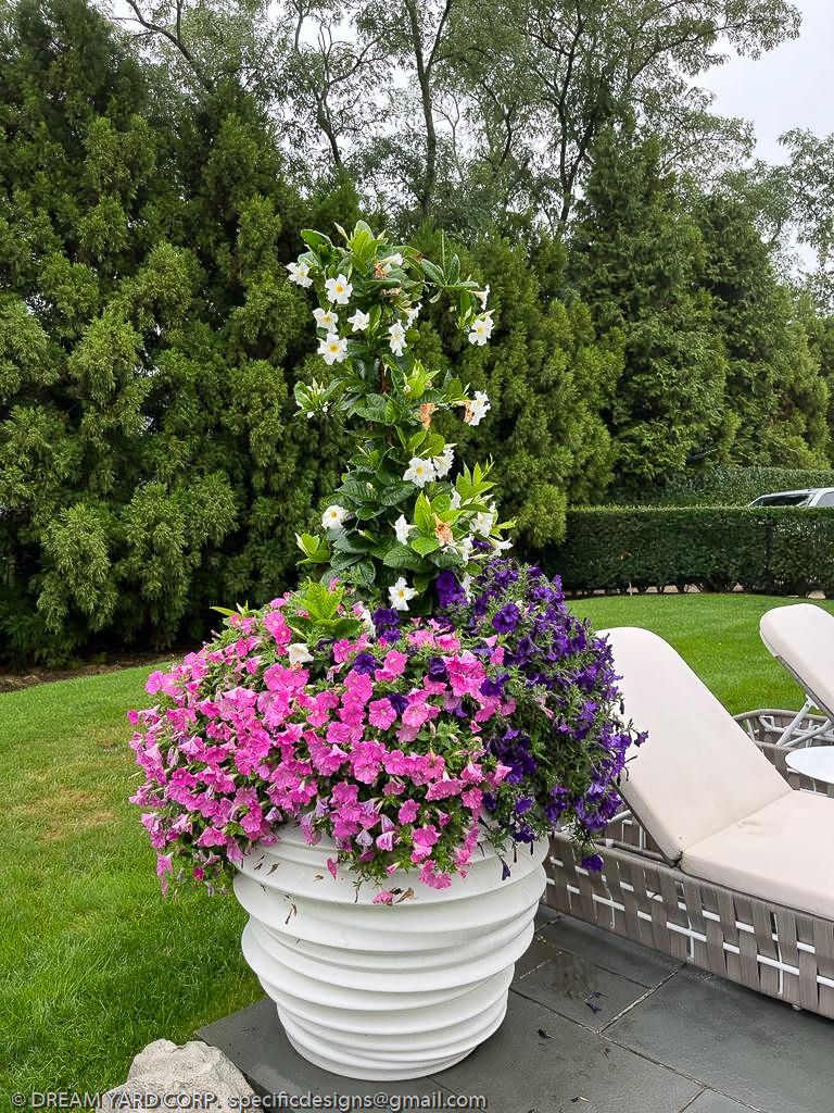 Large white planter overflowing with pink, purple, and white flowers on a lawn, with lounge chairs and trees in the background.