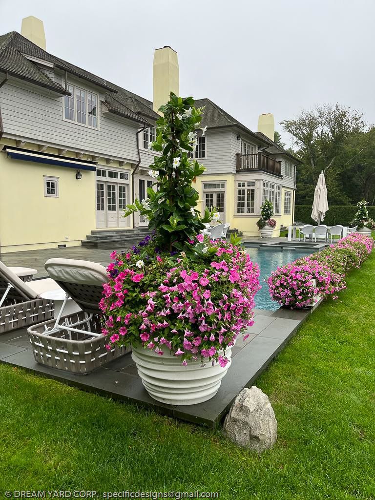 Large house with pool, flowerpots with pink petunias, lounge chairs, green grass.