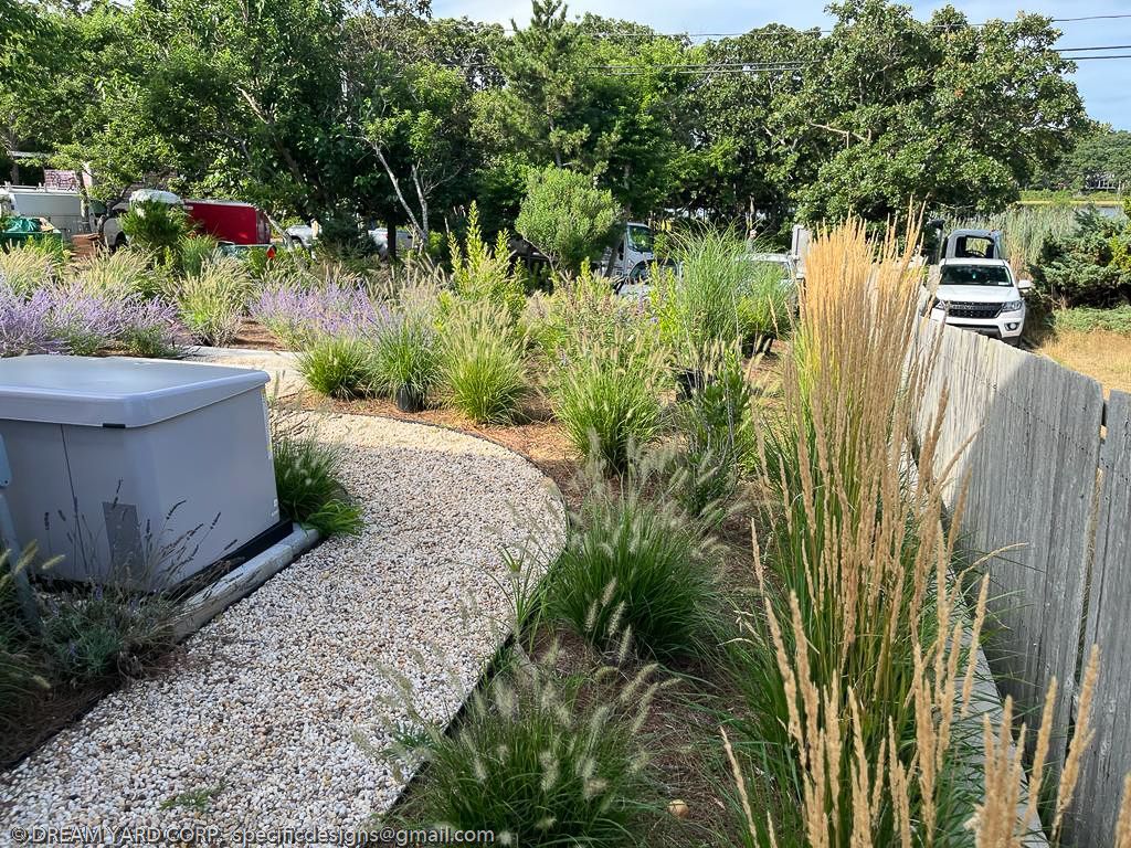 A landscaped garden with a winding gravel path, shrubs, and tall grasses. A grey utility box is on the left.
