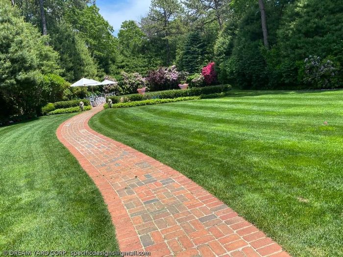 Brick path curves through green lawn, leading to garden with white structure and pink flowers.