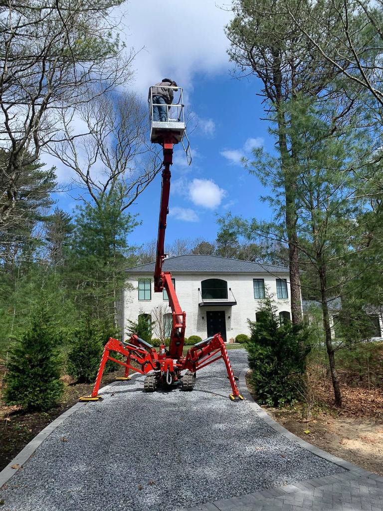 Red cherry picker extended toward the sky; person in the basket. Driveway, house, trees and blue sky in background.