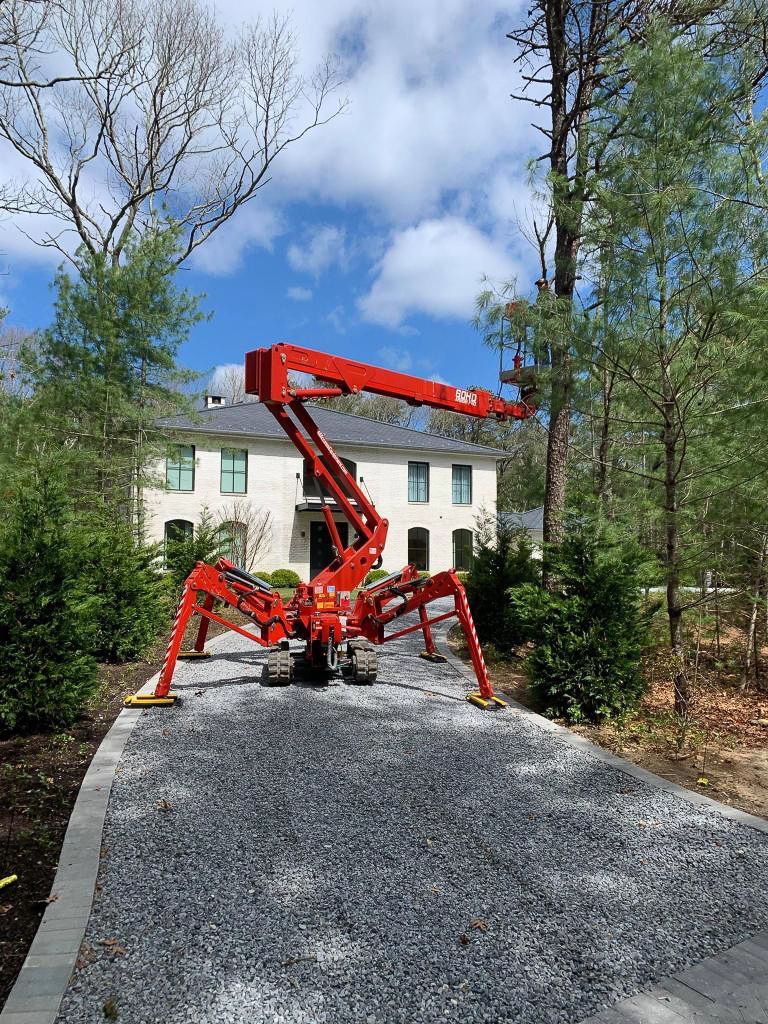 Red spider lift extended in front of a white house with a gravel driveway. Blue sky.