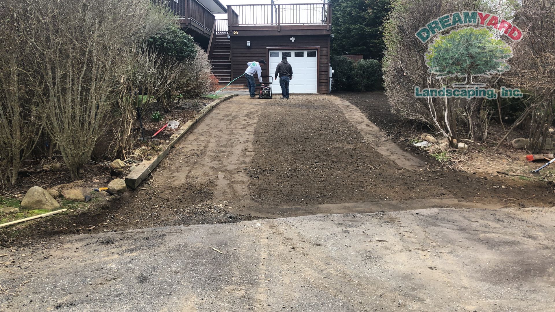 Two men working on a driveway, tilling the gravel. A garage and house are in the background.