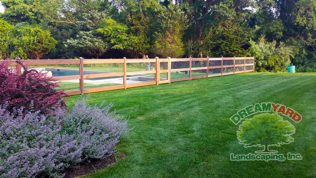 Wooden fence with mesh in a green yard, purple and green bushes in the foreground, trees in the background.