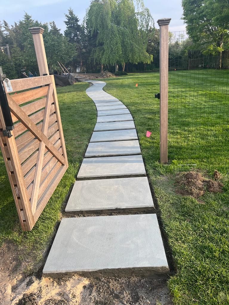 Stone path winding through green grass toward a wooden gate and fence, in a backyard setting.