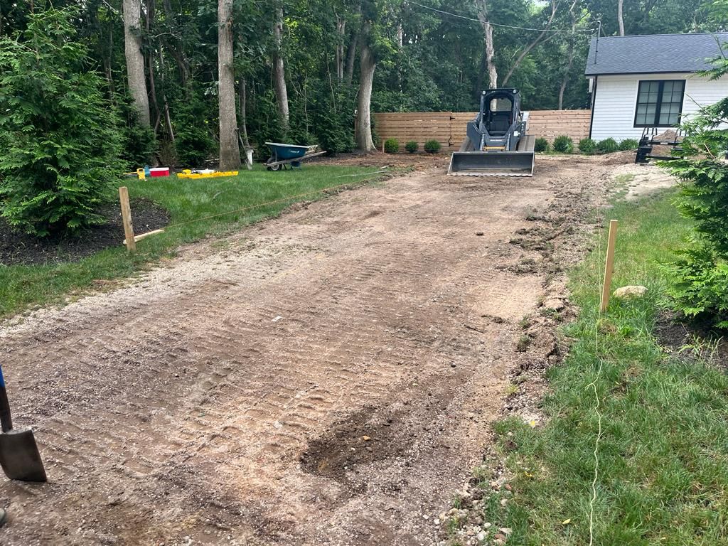A dirt driveway being graded by a small skid steer loader, beside green grass and a house.
