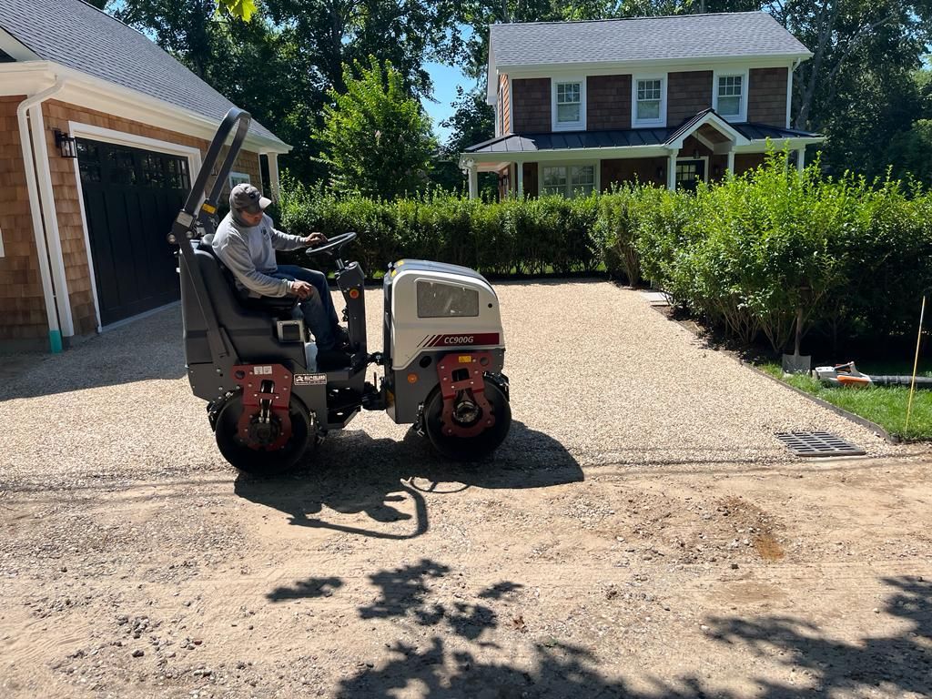 Man operating a road roller on a gravel driveway, house and garage in the background. Sunny day.