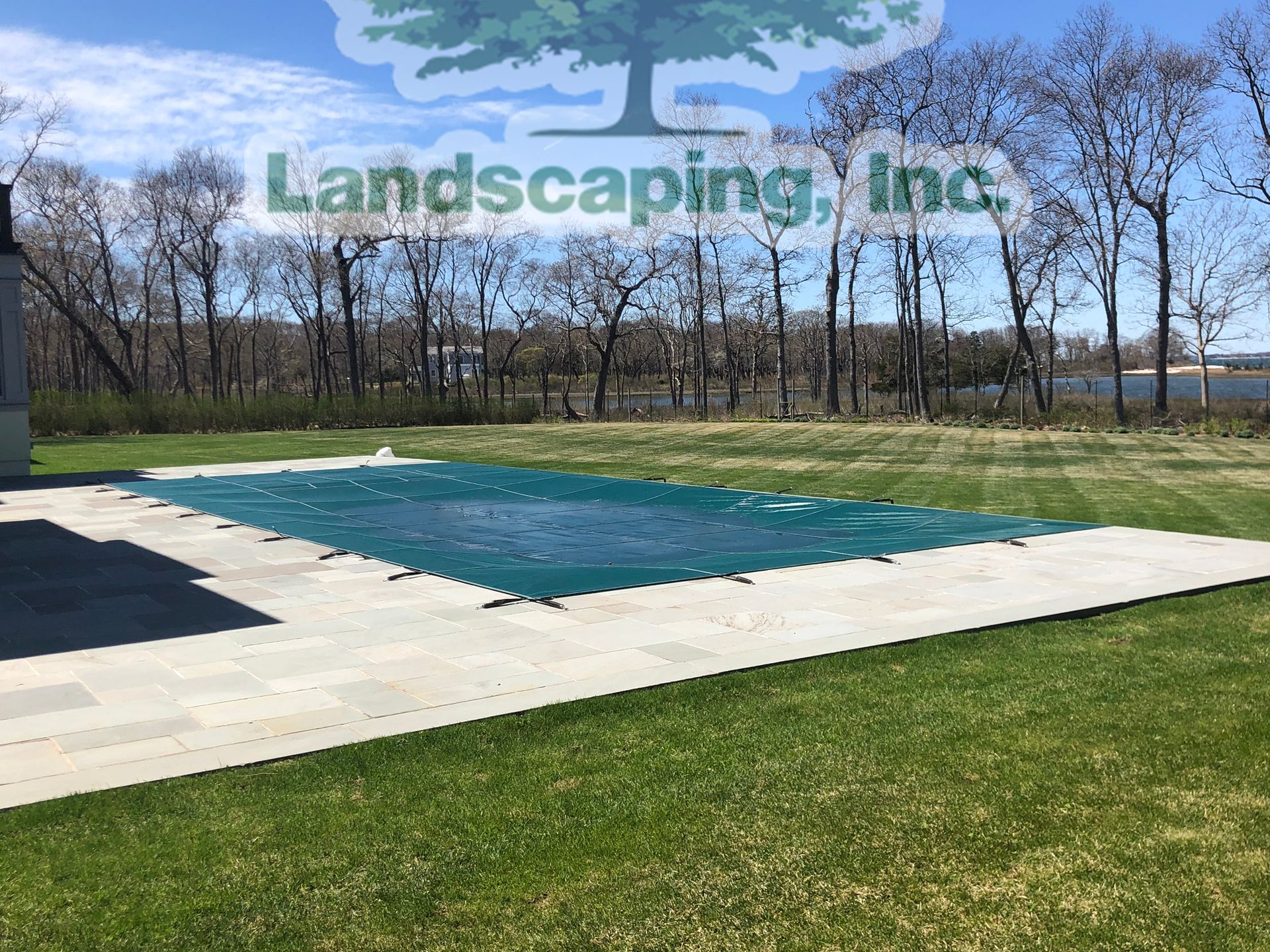 Pool covered by a green tarp, surrounded by a grassy lawn and a paved patio. Trees and sky in the background.