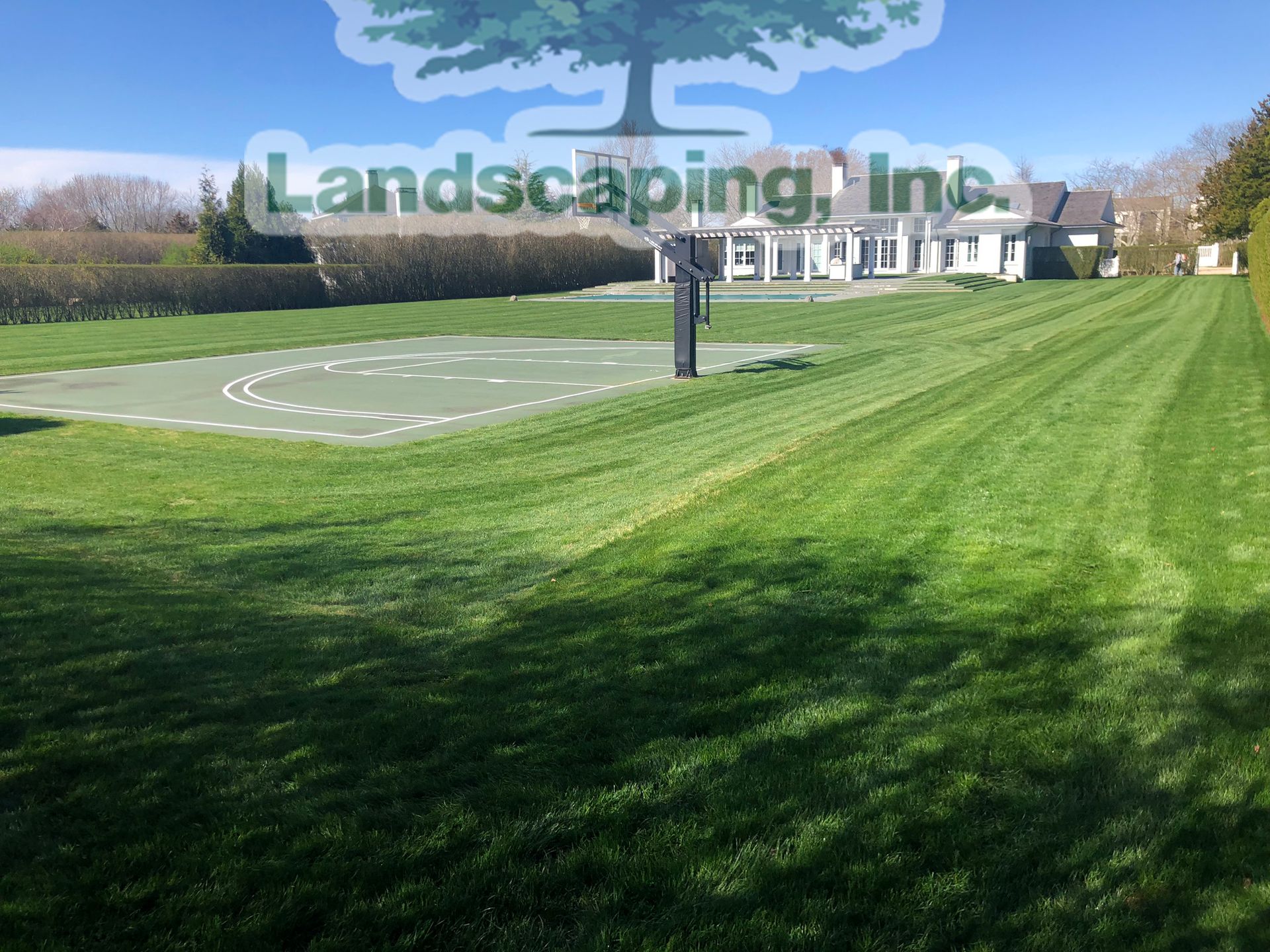 Well-manicured lawn with a basketball court and a white house in the background under a sunny sky.
