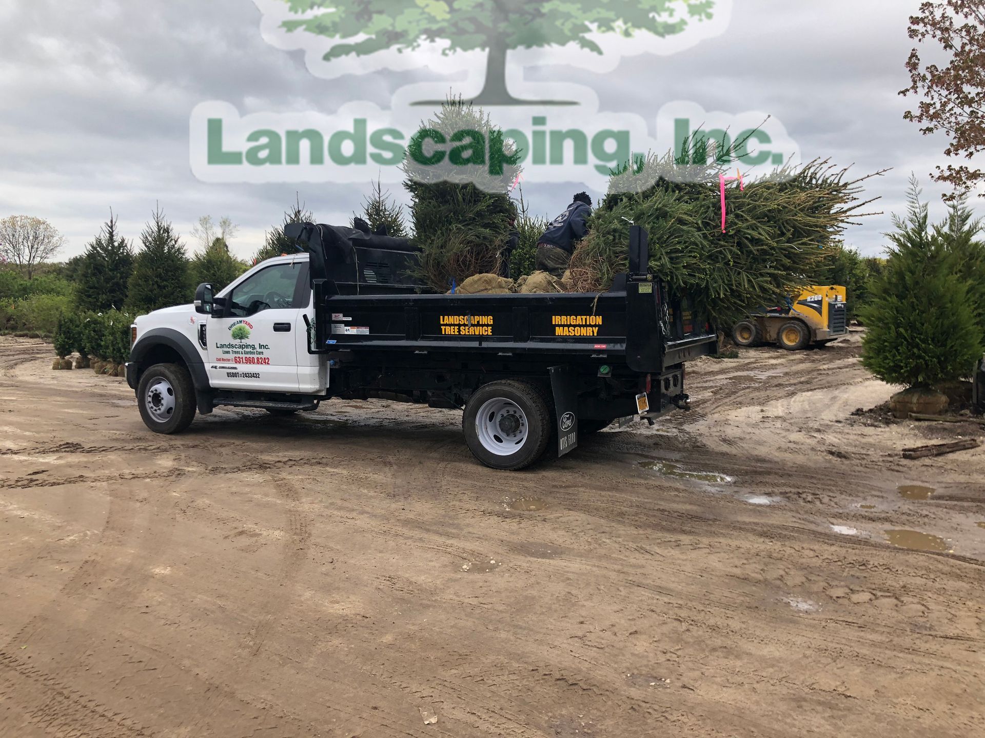 White landscaping truck with plants in the bed, on a dirt lot with other trees and a loader in the background.