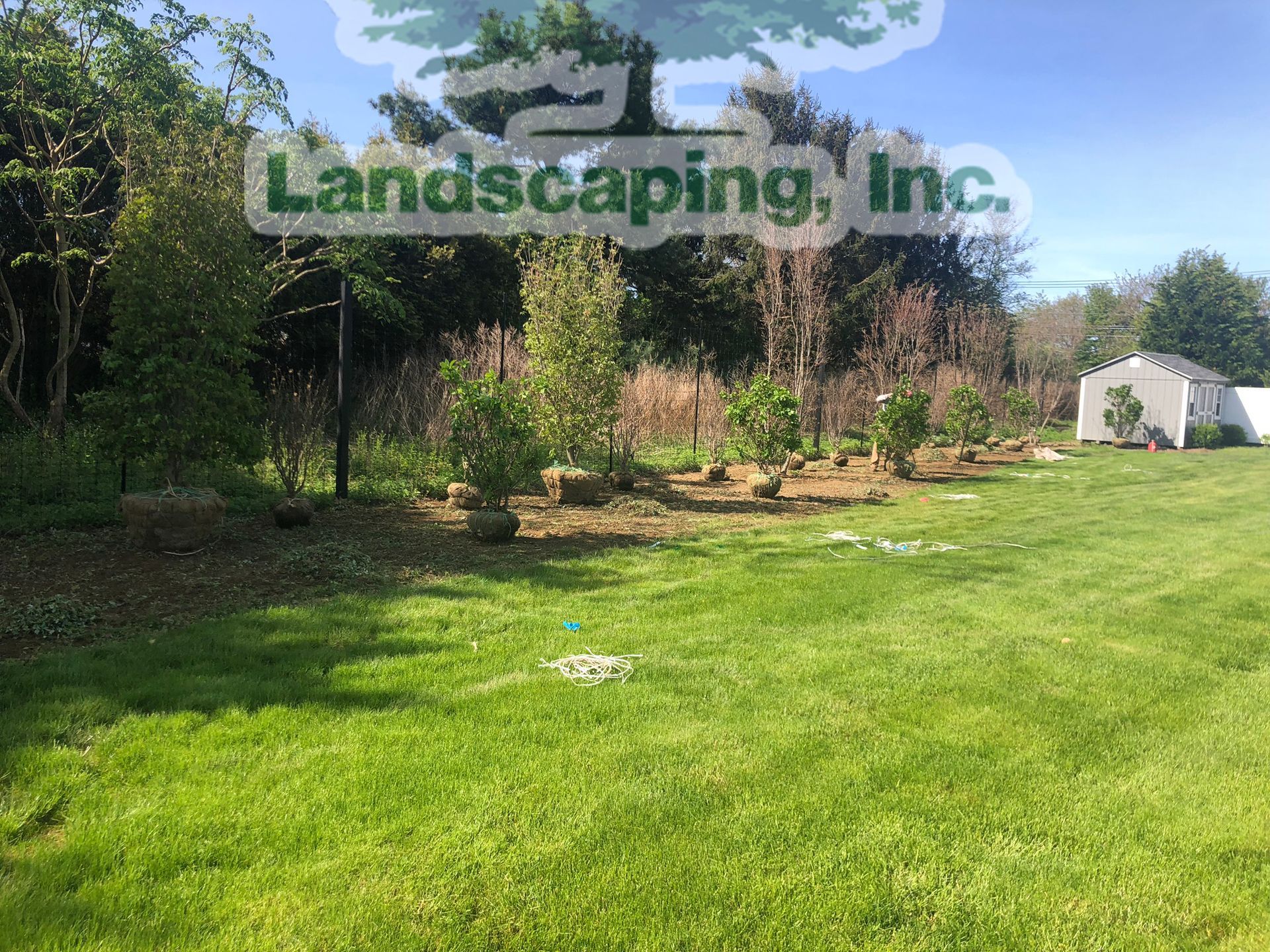 Lush green lawn with a small garden and shed, under a bright blue sky. Landscaping, Inc. logo.