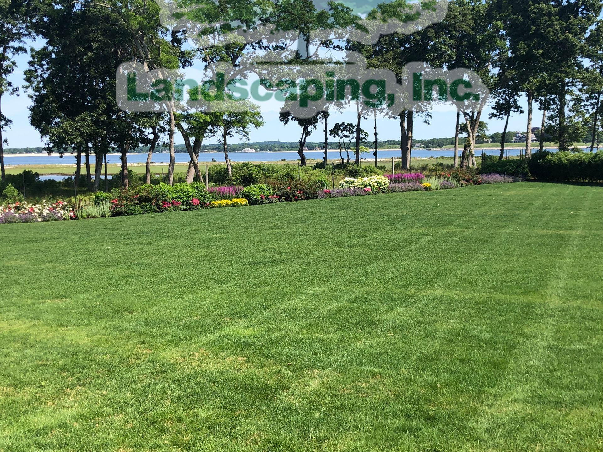 Lush green lawn with colorful flowerbeds overlooking a body of water lined with trees.