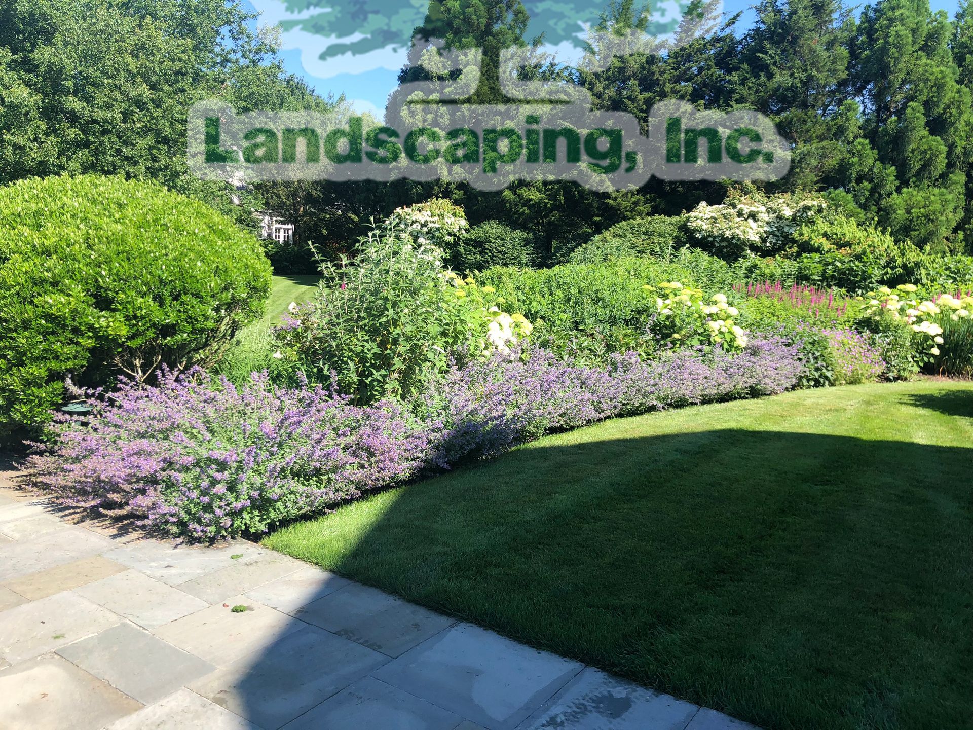 Green lawn and flower bed, next to stone patio; landscaping with purple flowers, green bushes, and trees.