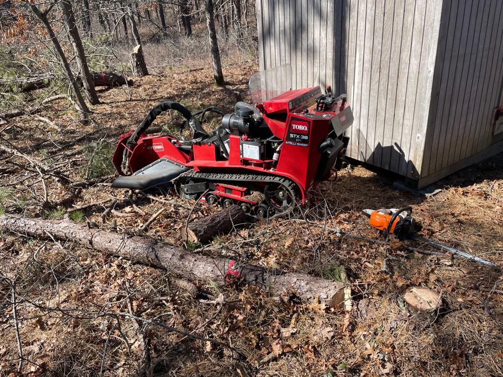 Red stump grinder next to a shed, wood debris, chainsaw on the ground. Outdoor setting.