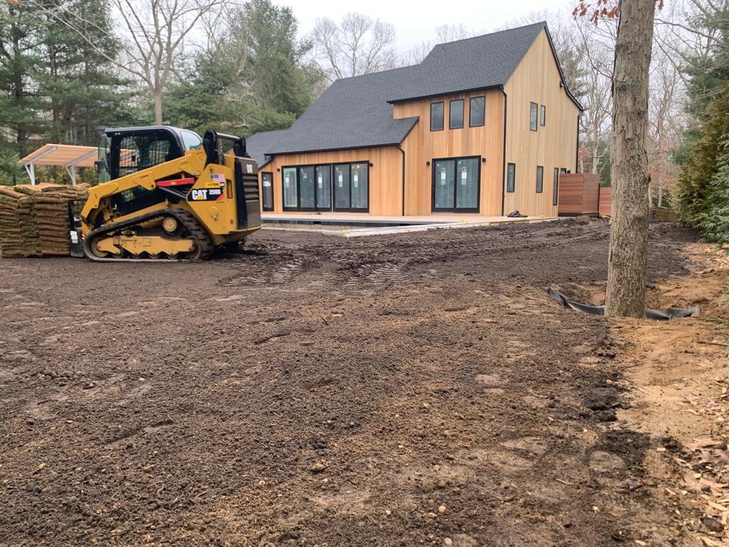 Yellow skid steer next to a modern wood house, grading soil.  Stacks of sod nearby.  Outdoor construction.