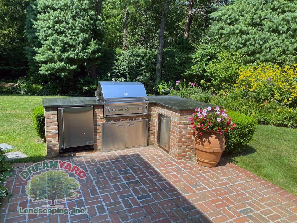 Brick outdoor kitchen with grill, refrigerator, and potted flowers on a brick patio surrounded by greenery.
