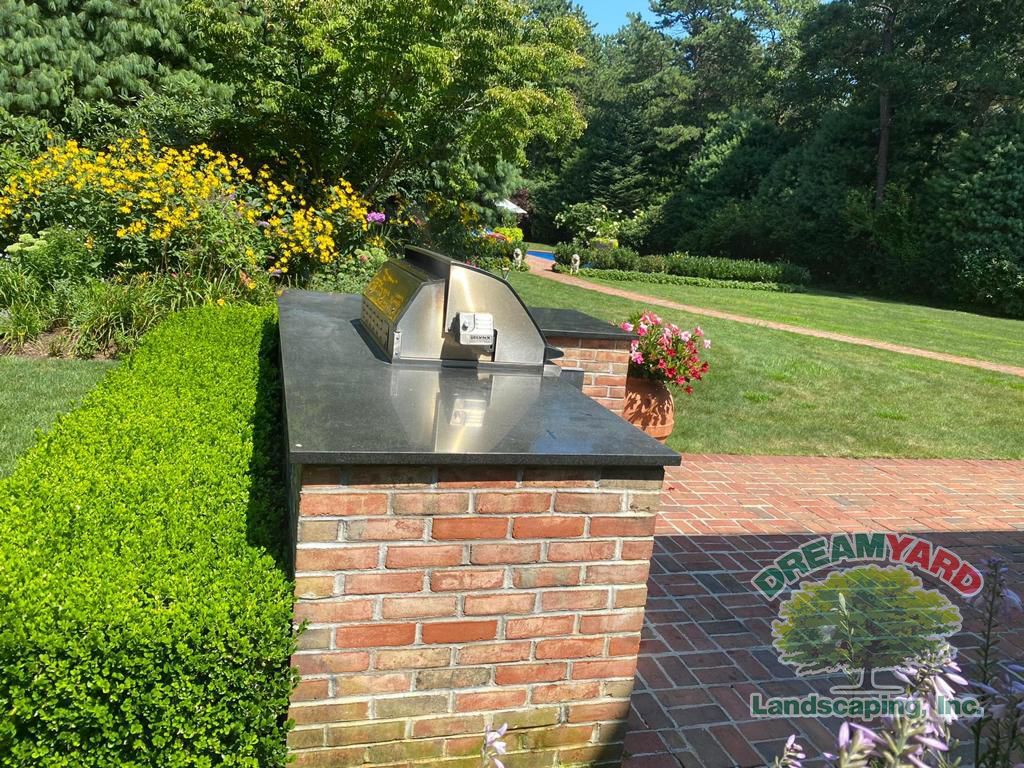 Brick outdoor kitchen with stainless steel grill, black countertop, and lush garden background.
