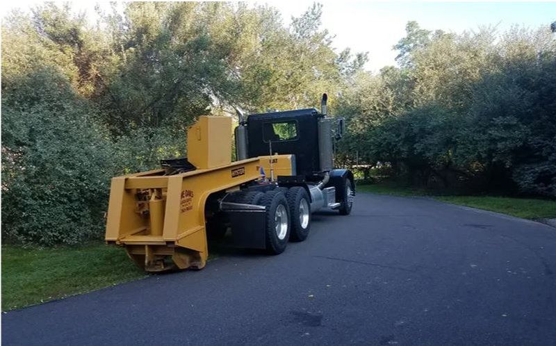 Yellow truck with a specialized trailer parked on a road, surrounded by trees.