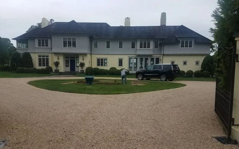 Large house with gravel driveway; a black SUV and two people stand near the entrance on an overcast day.