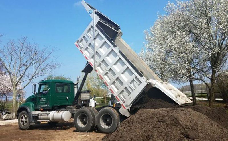 Green dump truck dumping mulch onto a brown pile on a sunny day.